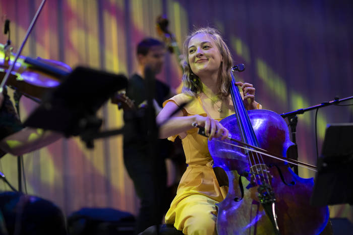 Alice, on stage in a concert, playing her cello whilst beaming at Patters