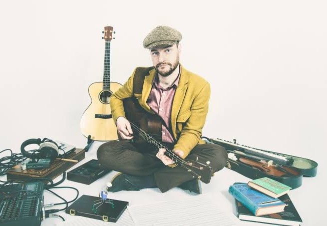 Mike, sat on the ground with his guitar in the playing position, surrounded by another guitar, books, a fiddle in it's case, and recording equipment